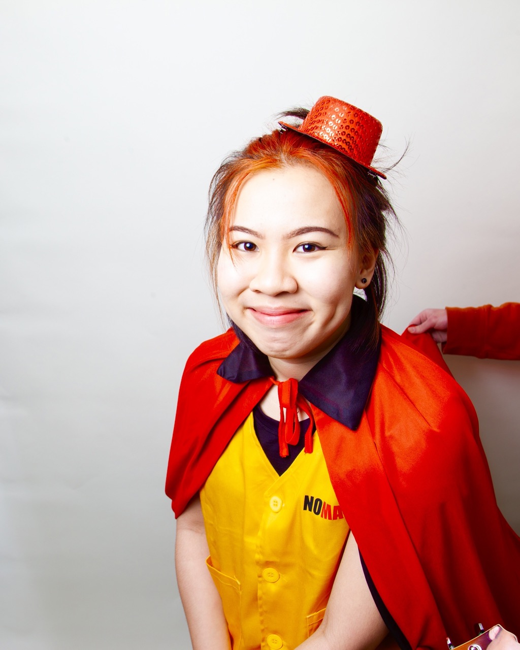 Kyra smiles at the camera wearing a yellow vest, red cape, and red bowler hat